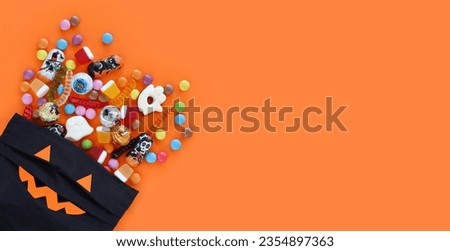 Halloween orange banner in corner with black textile bag with smiling pumpkin jack-o-lantern face. Pouch full of spilled assorted traditional candies. Trick-or-treating concept. With copy space.