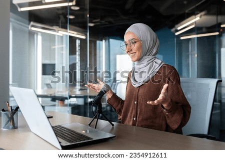 A smiling woman in a hijab sits in the office in front of a laptop and speaks into a microphone, records a podcast, conducts a meeting, an interview.