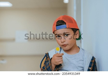 Little caucasian boy in baseball cap and plaid shirt  holds lollipop looks at camera with confident face expression with copy space on background. Cute male kid loves candy and sweets. Mockup.