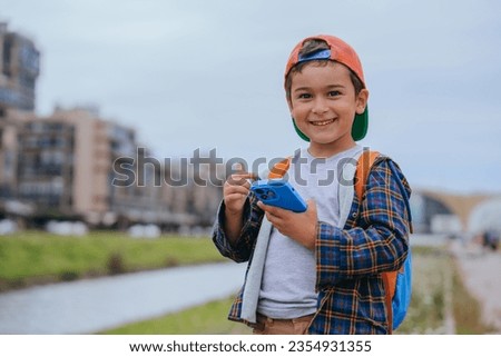 Cheerful little boy in casual dress with backpack holds phone smiles, enjoying games on summer sunny day outside. Preschooler kid excited by new phone. Childhood, technology, happy people.