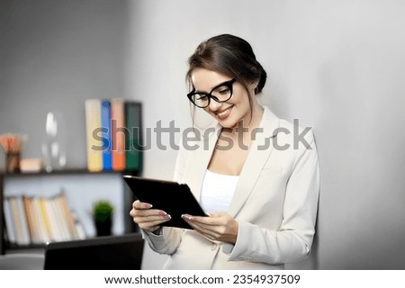 Attractive woman in black-framed glasses is smiling while reading or watching something on digital tablet at office or at home. Online shopping. Video call, online meeting. Modern technology concept.