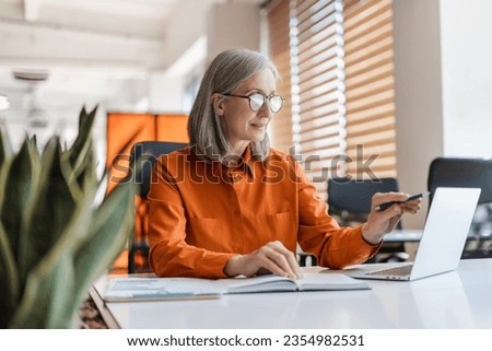 Portrait of beautiful confident senior woman, manager using laptop computer working online sitting in modern office. Business woman wearing stylish eyeglasses checking email. Technology concept 