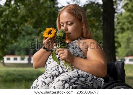 Smiling Happy Young Woman With Short Stature On Wheelchair Holds, Sniffing Flowers in Green Park At Summer Day. Female Adult With Disability. Copy Space For Text. Horizontal Plane. High quality photo Royalty-Free Stock Photo #2355036699