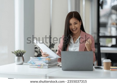 Cheerful and happy businesswoman working on laptop and financial documents in office, attractive smiling female office worker successful.