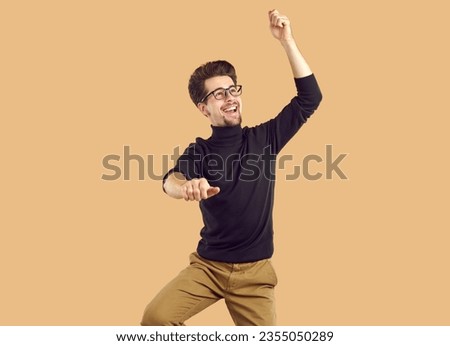 Happy attractive young man is dancing, having fun and fooling around. Bearded funny guy with glasses wearing black turtleneck and brown trousers on isolated beige background in the studio.