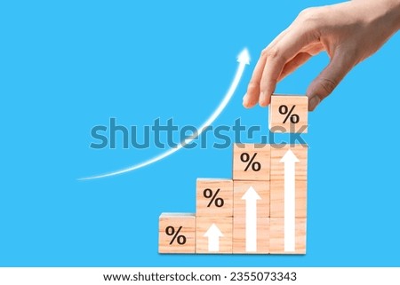 Hand stacking wooden blocks as symbol of increase with percentage symbol and arrow. concept of financial goals and interest rates on blue background.