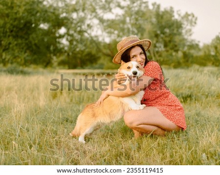 Beautiful girl with long black hair wearing a short red dress and a hat.  Corgi dog in the background of green grass. A girl is hugging her corgi dog. Walking the dog, friendship. Summer picture