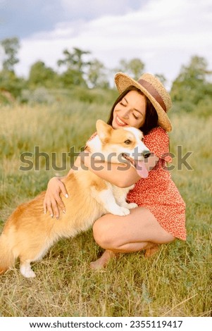 Beautiful girl with long black hair wearing a short red dress and a hat.  Corgi dog in the background of green grass. A girl is hugging her corgi dog. Walking the dog, friendship. Summer picture
