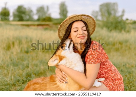 Beautiful girl with long black hair wearing in red dress and a hat.  Corgi dog in the background of green grass. A girl gently strokes her corgi dog. Dog is licking the gir, friendship. Summer picture