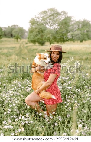 Beautiful girl with long black hair wearing a short red dress and a hat.  Corgi dog in the background of green grass. A girl is holding her corgi dog. Walking the dog, friendship. Summer picture