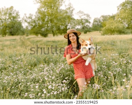 Beautiful girl with long black hair wearing a short red dress and a hat.  Corgi dog in the background of green grass. A girl is holding her corgi dog. Walking the dog, friendship. Summer picture