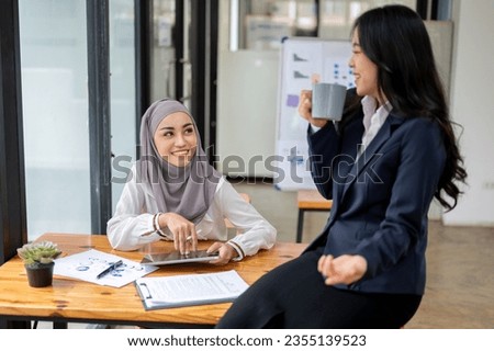 An attractive and smiling Asian Muslim female office worker wearing a hijab enjoys chatting with her colleague during a coffee break in the office. Royalty-Free Stock Photo #2355139523