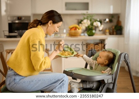 Mother spoon feeding her baby boy in the kitchen Royalty-Free Stock Photo #2355180247