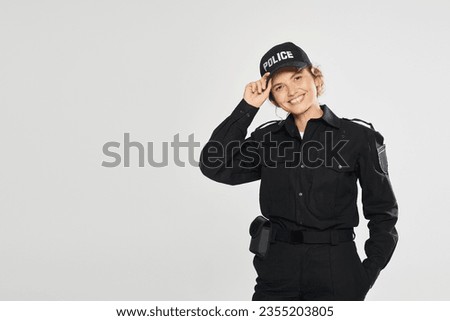cheerful policewoman in uniform touching cap and looking at camera isolated on grey Royalty-Free Stock Photo #2355203805