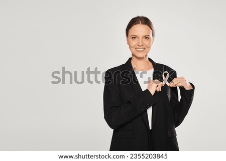 positive businesswoman in suit holding pink ribbon of breast cancer isolated on grey Royalty-Free Stock Photo #2355203845
