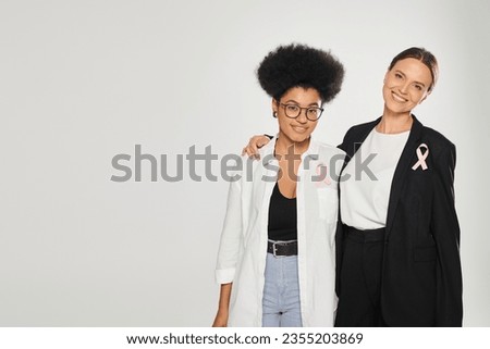 happy businesswoman with pink ribbon hugging african american woman isolated on grey, breast cancer Royalty-Free Stock Photo #2355203869