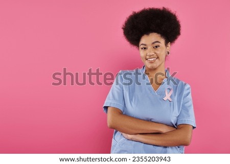 smiling african american doctor with ribbon crossing arms isolated on pink, breast cancer concept Royalty-Free Stock Photo #2355203943