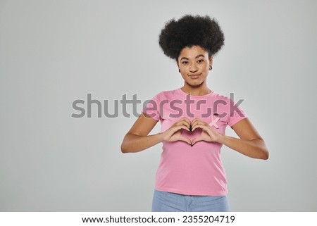 breast cancer awareness, african american woman with pink ribbon on grey backdrop, heart sign, smile Royalty-Free Stock Photo #2355204719