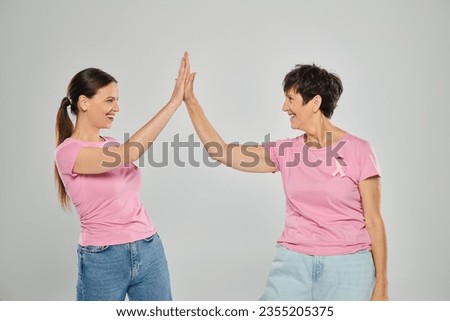 breast cancer concept, happy women with pink ribbons giving high five on grey backdrop, cancer free Royalty-Free Stock Photo #2355205375