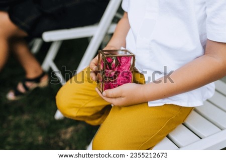A child, a boy, holds in his hands a glass box with flowers, golden rings with words of love forever at the wedding ceremony. Photography, portrait.