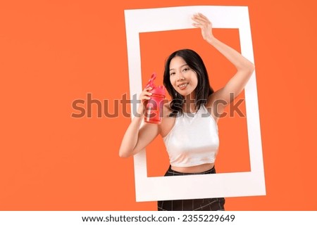 Young beautiful Asian woman with frame and bottle of water on orange background