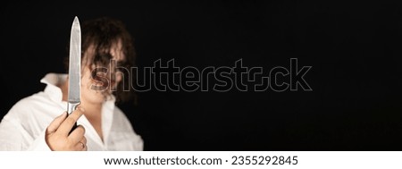 Halloween banner. A man with a knife on a black background in the form of a crazy killer clown looks at the camera. Copy space. Scary clown smiling at the camera holding a knife in front of him.