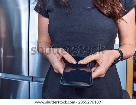 Woman holding phone in black t-shirt in kitchen. Long brown hair. Smart watch on hands