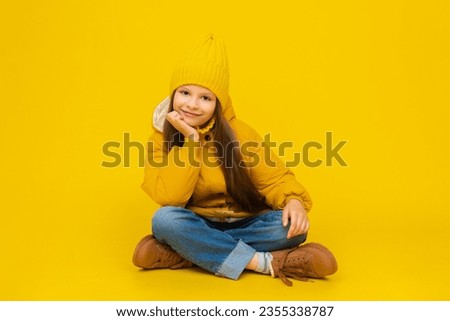 A young girl is sitting on the floor in full height, cross-legged, in autumn warm clothes. A child in a yellow warm jacket and a long beanie hat. Clothes for children. Yellow isolated background.