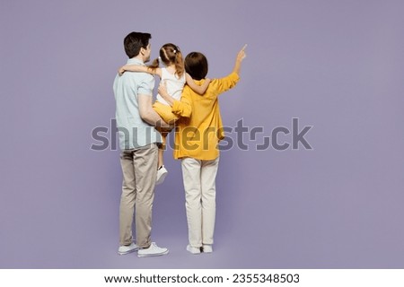Full body back rear view young parents mom dad child kid girl 6 years old wearing blue yellow casual clothes hold daughter point finger aside isolated on plain purple background. Family day concept Royalty-Free Stock Photo #2355348503