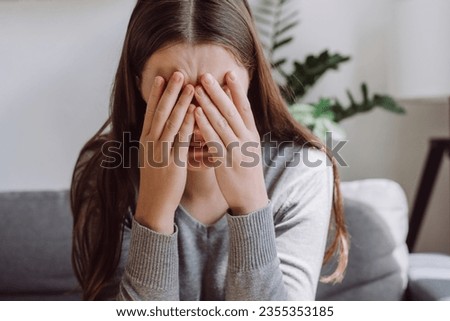 Lost in thoughts stressed brunette female regretting of wrong decision. Close up of unhappy anxiety young caucasian female covering face with hands sitting on grey couch in living room at home Royalty-Free Stock Photo #2355353185