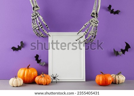 Embracing the spooky spirit of Halloween. Side view photo of scary skeleton hands, pumpkins, creepy spiders and bat silhouettes on table with purple background and empty frame for promo or text