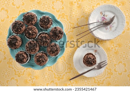 Mini chocolate cupcakes on turquoise cakestand with small plates and rose gold forks on yellow patterned tablecloth.  Shot from above in flat lay composition in horizontal format. Royalty-Free Stock Photo #2355421467