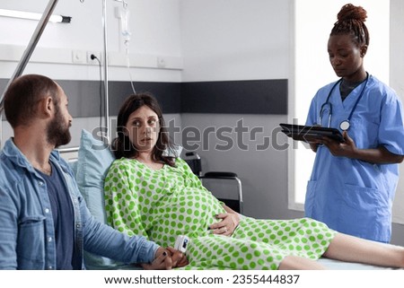 Couple receiving medical assistance from african american nurse before childbirh in hospital ward. Nurse typing woman contractions level on tablet computer, explaining pregnancy labor process