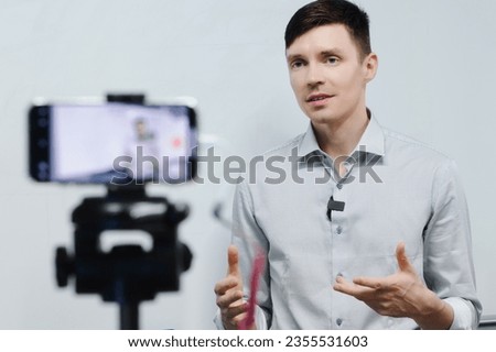 Young guy records video blog on phone camera indoors. Caucasian man conducts training remotely. Standing in front of phone. Live broadcast or streaming.