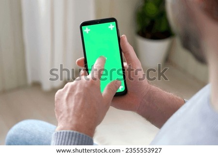 Young man sitting at home holding smartphone green mock-up screen in hand. Male person using chroma key mobile phone. Vertical mode. Touching, swiping display, tapping, surfing Internet social media