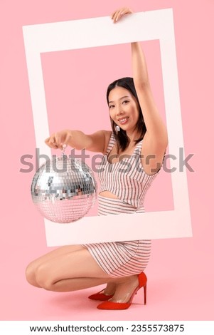 Young beautiful Asian woman with frame and disco ball sitting on pink background
