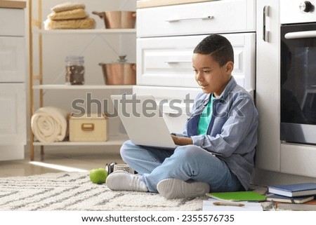 Little African-American boy with laptop studying computer sciences online in kitchen Royalty-Free Stock Photo #2355576735