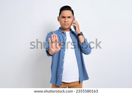 Serious young Asian man in casual shirt talking on smartphone and making stop sign with hand isolated on white background