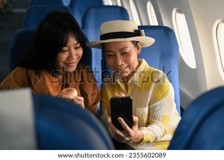 Happy middle aged woman and daughter sitting in passenger airplane and taking picture, waiting for airplane landing