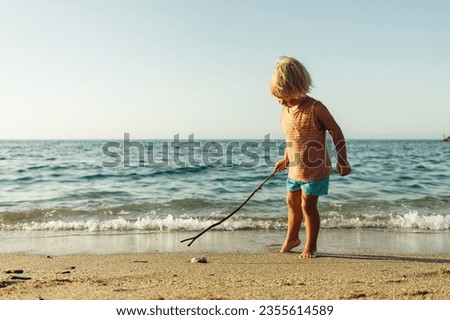 a little blond boy walks along the seashore with a stick in his 