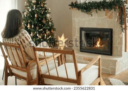Woman in cozy sweater relaxing on modern chair and looking at fireplace with festive mantle on background of stylish decorated christmas tree with lights. Winter holidays