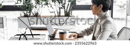 side view of experienced middle aged manager working on laptop near coffee to go in office, banner
