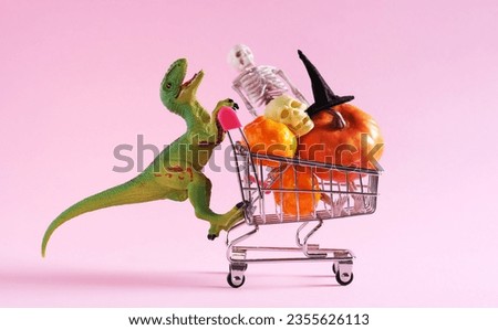 Happy green dinosaur with shopping trolley full of Halloween decorations on pink background.