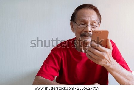 Portrait picture of Asian old man sitting on chair and holding, looking at smart phone screen at home. Retirement age concept.