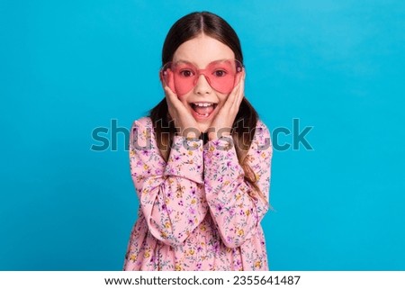 Photo of cute little girl touch cheeks excited heart sunglass dressed stylish pink flower print clothes isolated on blue color background