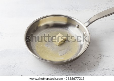 A piece of butter is melted in a frying pan on a light gray background. Making cheese pasta sauce, step by step, do it yourself, step 1 Royalty-Free Stock Photo #2355677315