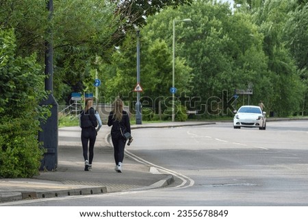 Urban girls in black outfits walking on sidewalk near road, carrying bags. Lined with trees, bushes, road signs, street lights, and white car passing by.