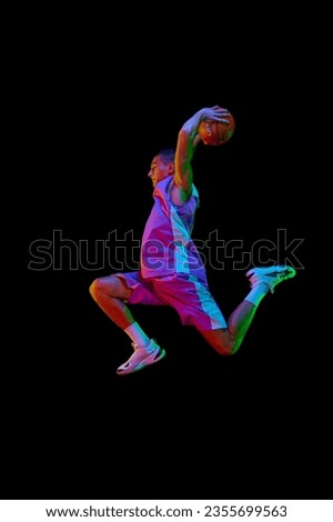 Scoring winning goal. Young man in uniform playing basketball, jumping with ball against black studio background in neon light. Concept of professional sport, competition, hobby, game, competition
