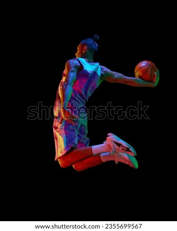 Athletic young guy, basketball player in motion during match, jumping with ball against black studio background in neon light. Concept of professional sport, competition, hobby, game, competition
