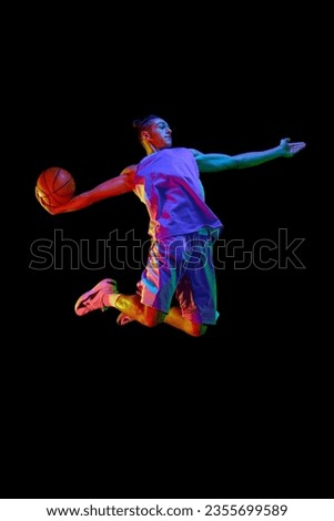 Dynamic image of young man, basketball player in uniform in motion, jumping with ball over black studio background in neon light. Concept of professional sport, competition, hobby, game, competition